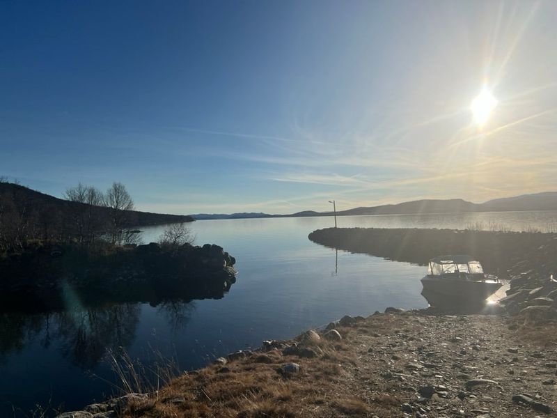 Sola lavt over Møsvatn sett fra strandkanten ved Galar, med båter fortøyd innenfor moloen