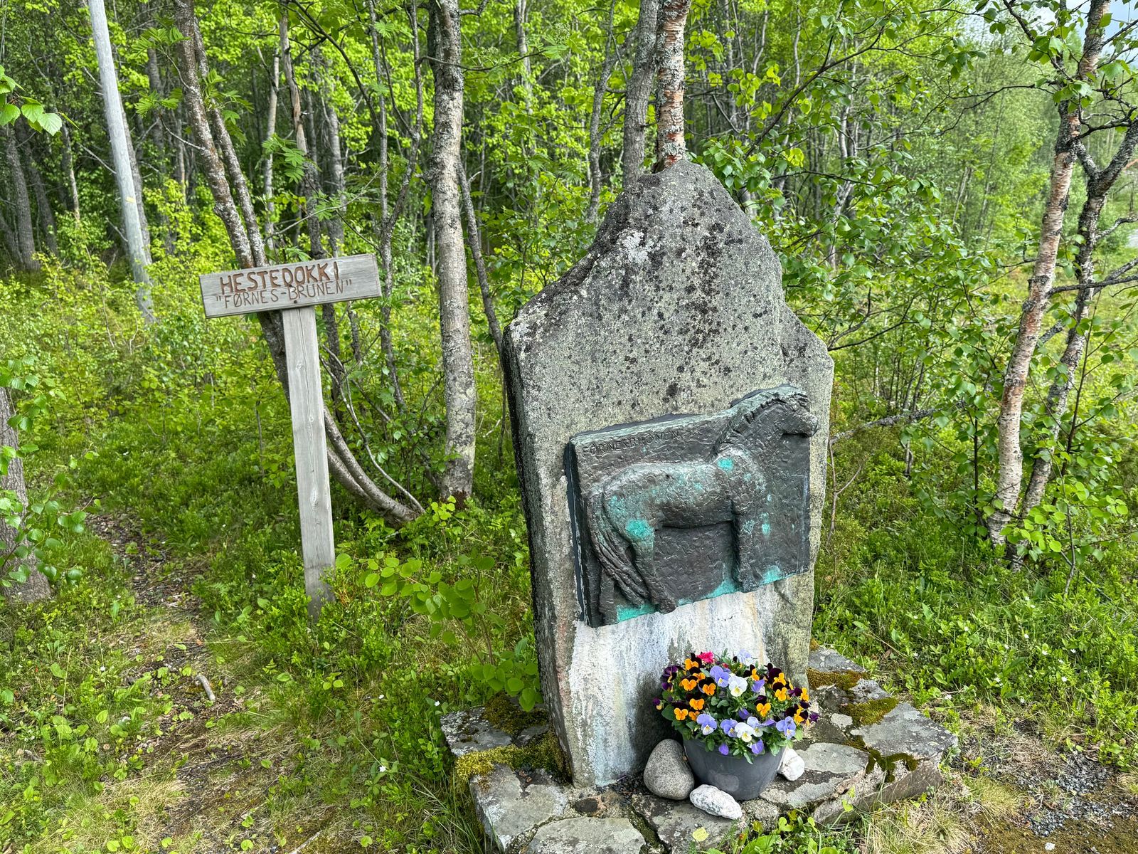 Minnestein over Førnesbrunen i bjørkeskogen, med en relieff av hesten og blomster ved foten