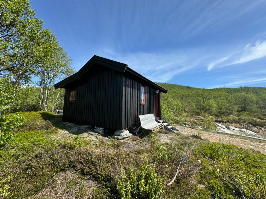 Bonserud, en mørk hytte på en grønn bakke med Møsvatn og fjellene i bakgrunnen
