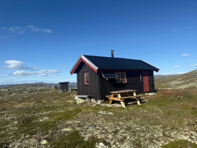 Jutulsbu med rødt vindsperre og bord utenfor, på steinete vidde under blå himmel