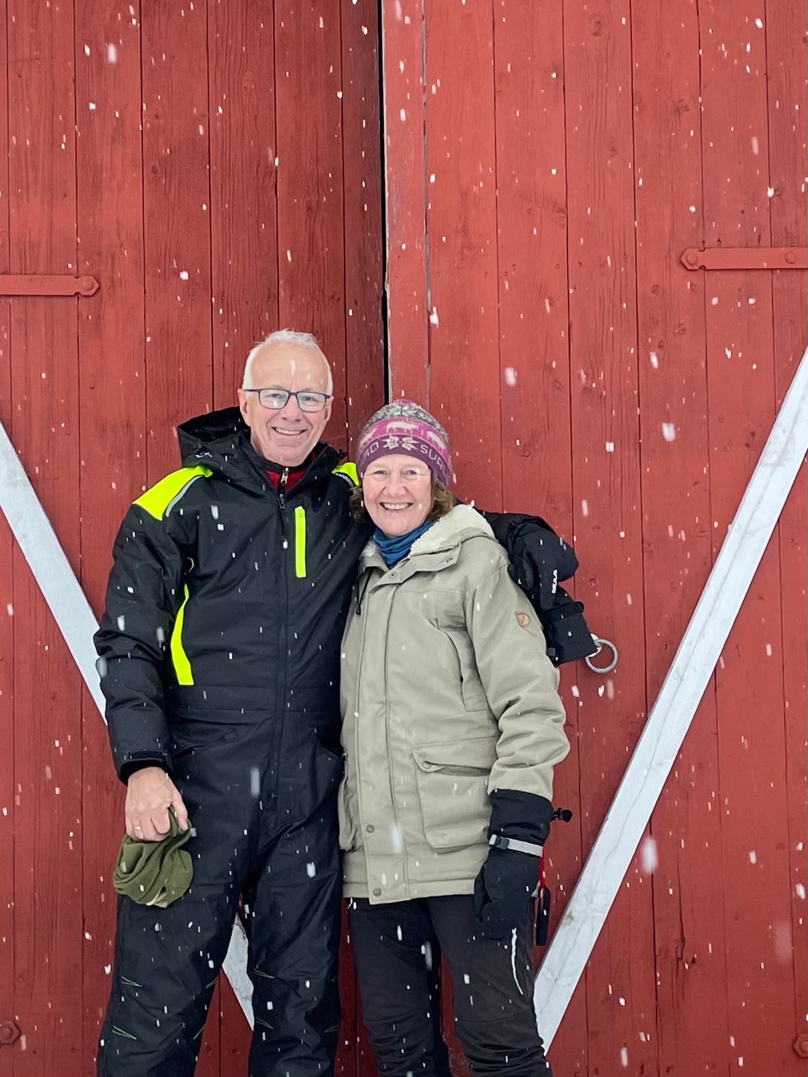 Mai og Knut Gunnarsli i vinterklær foran en rød låvedør på Førnes mens snøen faller