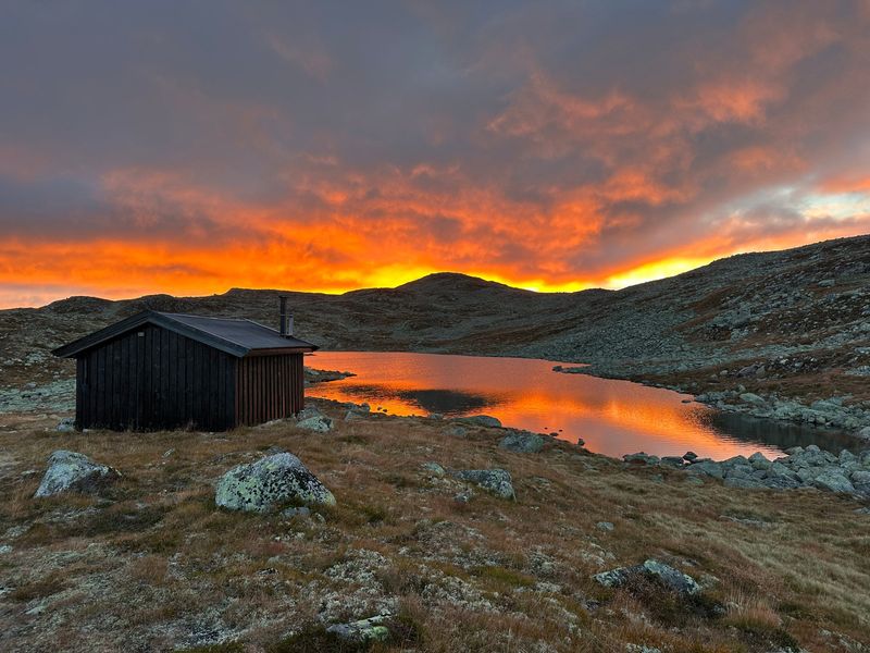 Mørk tømmerhytte ved et stille tjern på vidda under en brennende oransje soloppgang