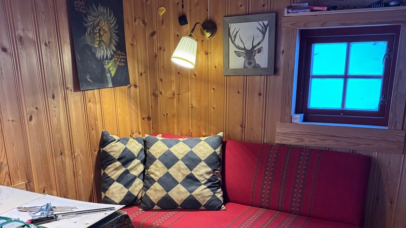 Inne i Såtehytta: rød sofa med puter langs tømmerveggen, lampe og vindu med blå himmel utenfor
